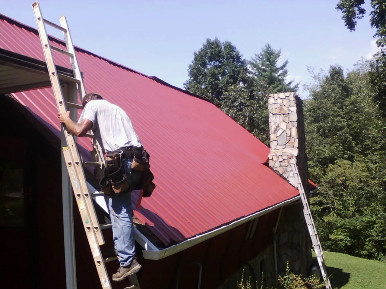 Roofer on a ladder beside a red metal roof and stone chimney — Soffit & Fascia Repair by Roofing Brothers in Joliet